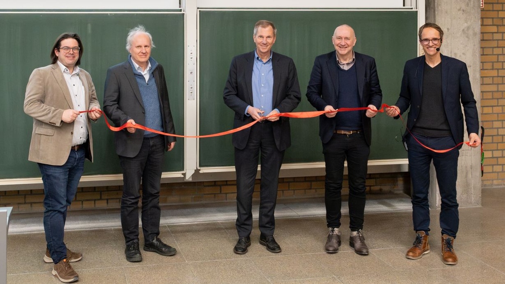 Five men in suits holding a red ribbon in front of a large chalkboard.